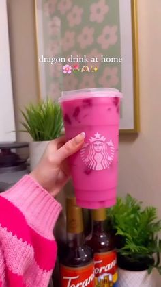 a person holding a pink starbucks cup in front of a potted plant and other items