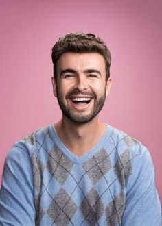 a man with a beard smiling and wearing a blue sweater against a pink background,