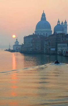 the sun is setting over some buildings on the water with a boat in front of it