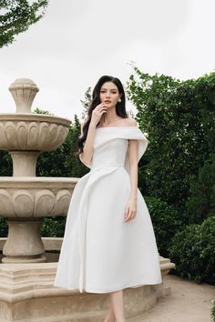 a woman wearing a white dress standing in front of a fountain with her hand on her face