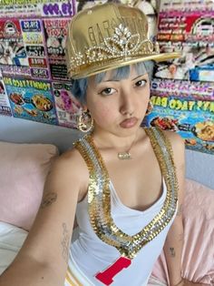 a woman with blue hair wearing a gold crown and necklaces on her head, sitting on a bed