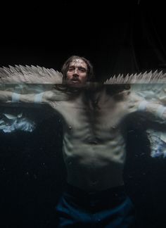 a man with white wings floating in the dark water, looking down at his shirtless torso