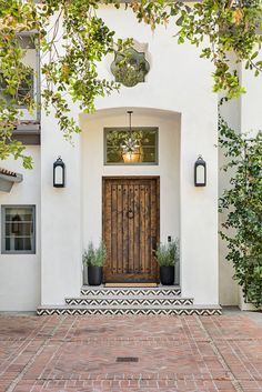 a white house with a wooden door surrounded by greenery and potted plants on the front porch