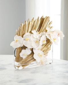 white orchids and gold sticks in a clear vase on a marble countertop with sunlight streaming through the window