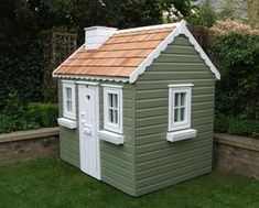 a small green house sitting in the grass next to a fence and shrubbery with white windows