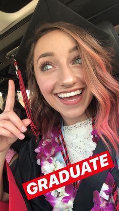 a woman with pink hair wearing a graduation gown and holding up a red sign that says graduate