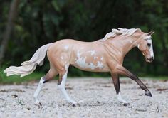 a toy horse that is walking in the sand with trees in the background and grass on the ground