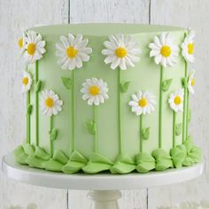 a green cake decorated with daisies and leaves on a white pedestal next to flowers