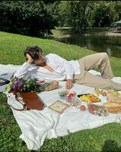 a man laying on top of a white blanket next to a river and picnicking