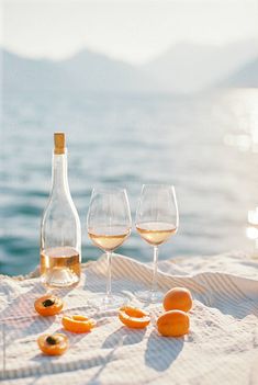 two wine glasses and three oranges on a towel near the water with mountains in the background