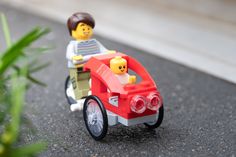 a lego man riding on the back of a red toy car next to a green plant