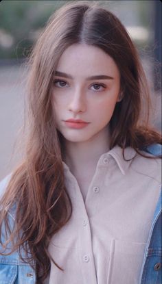 a young woman with long hair wearing a denim shirt and looking at the camera while standing in front of a building