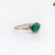 This elegant ring features a captivating 1.72 carat oval-shaped east west emerald, set in a polished band. Flanking the central stone are two round natural earth mined diamonds, adding a touch of sparkle. The emerald's rich green color is beautifully complemented by the warm tone of the band, creating a harmonious and refined look. The design is understated yet sophisticated, focusing on the natural beauty of the gemstones. This ring is made with solid 14K Gold and natural Earth mined SI / G-H d Oval Emerald Ring With Bezel Setting, East West Emerald Ring, Emerald Set, Green Emerald Ring, Warm Tone, Jewelry Appraisal, Solitaire Studs, Rich Green, Elegant Ring