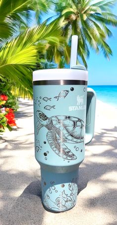 a blue coffee cup sitting on top of a sandy beach next to the ocean and palm trees