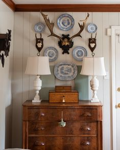 an antique dresser with blue and white plates mounted on the wall above it, along with deer heads