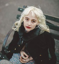 a woman sitting on top of a wooden bench next to a park bench with her hands in her pockets