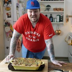 a man in a red shirt and blue hat standing next to a pan of food