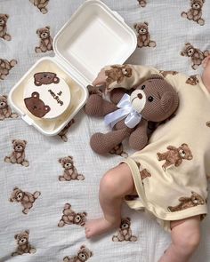 a baby laying in bed next to a brown teddy bear and a white tray with food