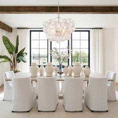 a dining room table with white chairs and vases on top of it, in front of a large window