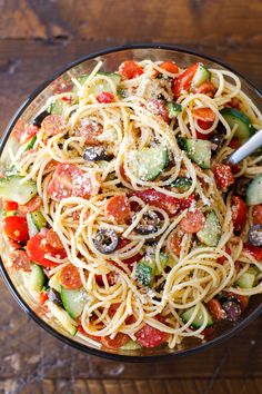 a glass bowl filled with pasta and vegetables on top of a wooden table next to a fork