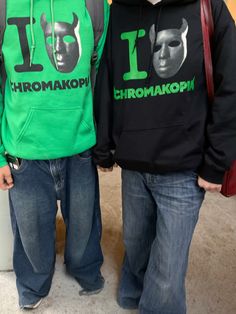 two men standing next to each other in front of a wall wearing green and black hoodies