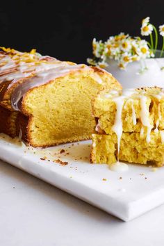 a loaf of cake with icing sitting on a white plate next to some flowers