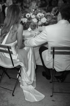 a man and woman sitting at a table with flowers in their hair, dressed in white