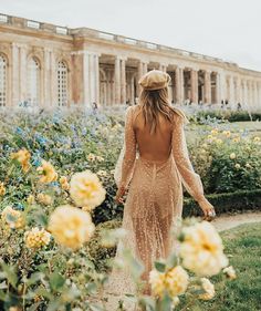 a woman in a dress and hat walking through flowers with a building in the background