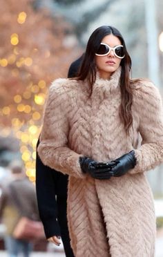 a woman in a fur coat and black gloves walking down the street with her hand on her hip