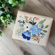 an embroidered box with flowers on it next to a potted plant and green leaves