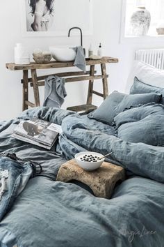 an unmade bed with blue linens in a white and gray bedroom, next to a wooden table