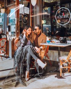 a man and woman sitting at a table in front of a store window talking on the phone