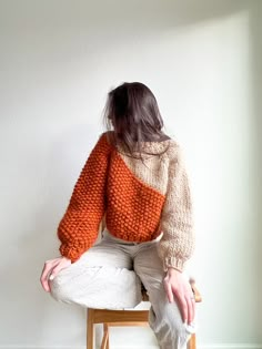 a woman sitting on top of a wooden chair wearing an orange sweater and white pants