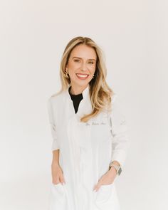 a woman standing in front of a white background wearing a white coat and black shirt