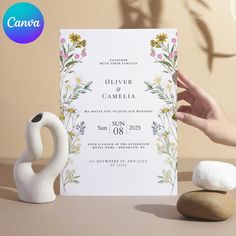 a person holding up a wedding card next to two rocks and a vase with flowers on it