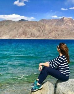 a woman sitting on top of a rock next to the ocean with mountains in the background