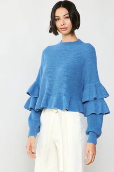 a woman wearing a blue sweater with ruffles on the sleeves and bottom, in front of a white background