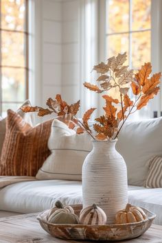 a white vase filled with leaves on top of a wooden table next to pillows and pumpkins