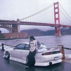 a woman sitting on the hood of a white sports car in front of a bridge