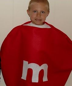 a young boy wearing a red cape with the letter m on it, standing in front of a white wall