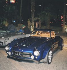 two classic cars parked next to each other on a brick road at night with palm trees in the background