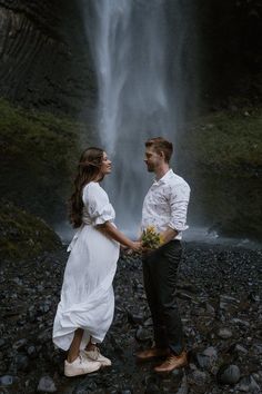 a man and woman standing in front of a waterfall with flowers on their hands, looking at each other