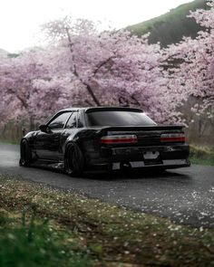 a black car parked on the side of a road next to trees with pink flowers