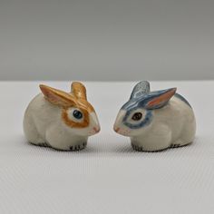 two ceramic rabbits sitting next to each other on a white tableclothed surface, one has blue eyes and the other has orange ears