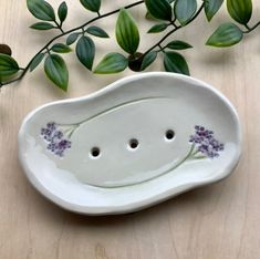 a ceramic bowl with two holes in the middle and purple flowers on it, next to some green leaves