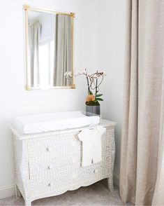 a white dresser with a mirror and flowers on it in front of a curtained window