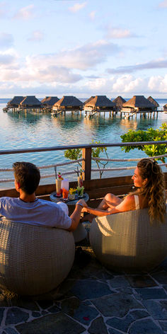 two people sitting on bean bag chairs in front of the ocean with thatched huts