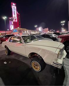 an old white car parked in a parking lot next to other cars and neon signs