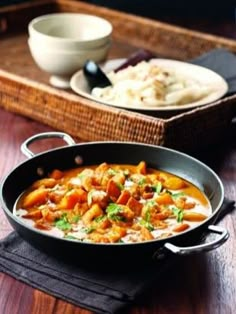 a pan filled with food sitting on top of a wooden table next to a bowl of rice