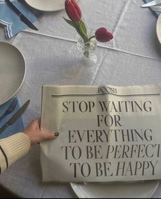 a person is holding up a newspaper on a table with plates and flowers in the background